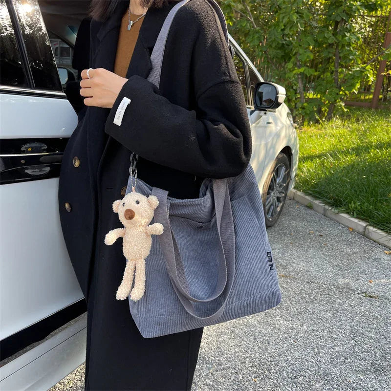 Woman wearing black coat holding grey corduroy tote bag with teddy bear keychain outdoors near white car