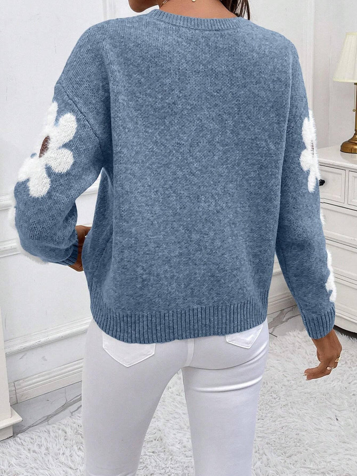 Back view of woman wearing blue knit sweater with white flower patterns on sleeves and white pants indoors
