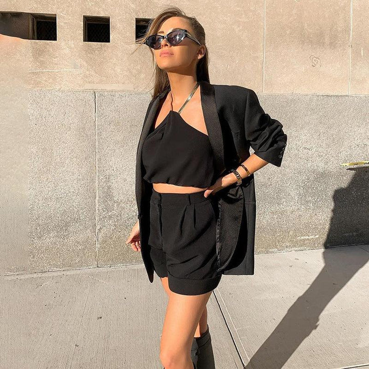Woman wearing black blazer, halter crop top, shorts, and sunglasses standing outdoors against stone wall