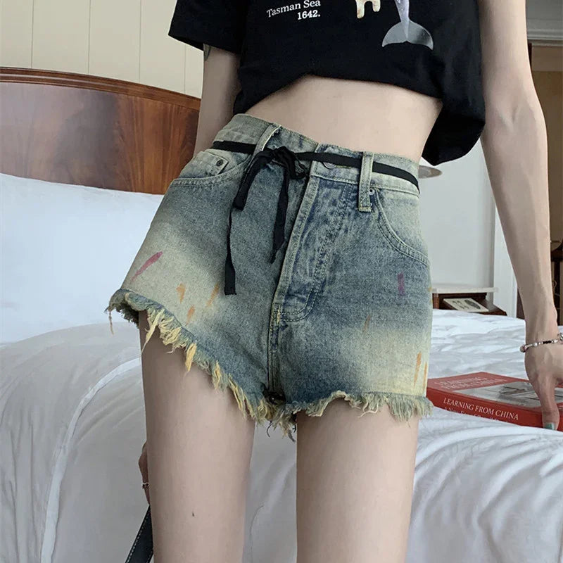 Woman wearing frayed high-waisted denim shorts with paint splatters and black belt, standing by bed