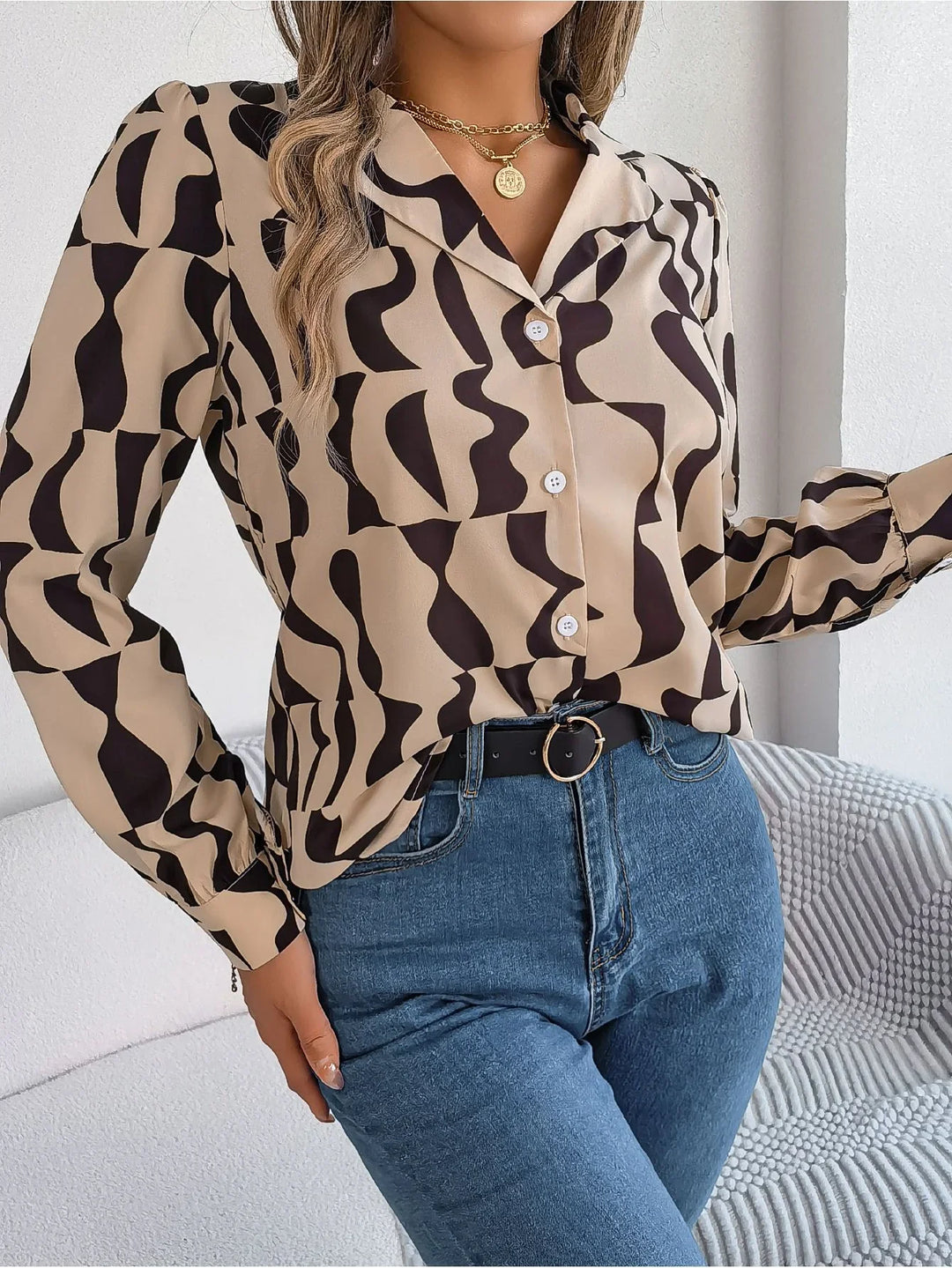 Woman wearing beige blouse with black abstract print and blue jeans, accessorized with gold necklace and black belt
