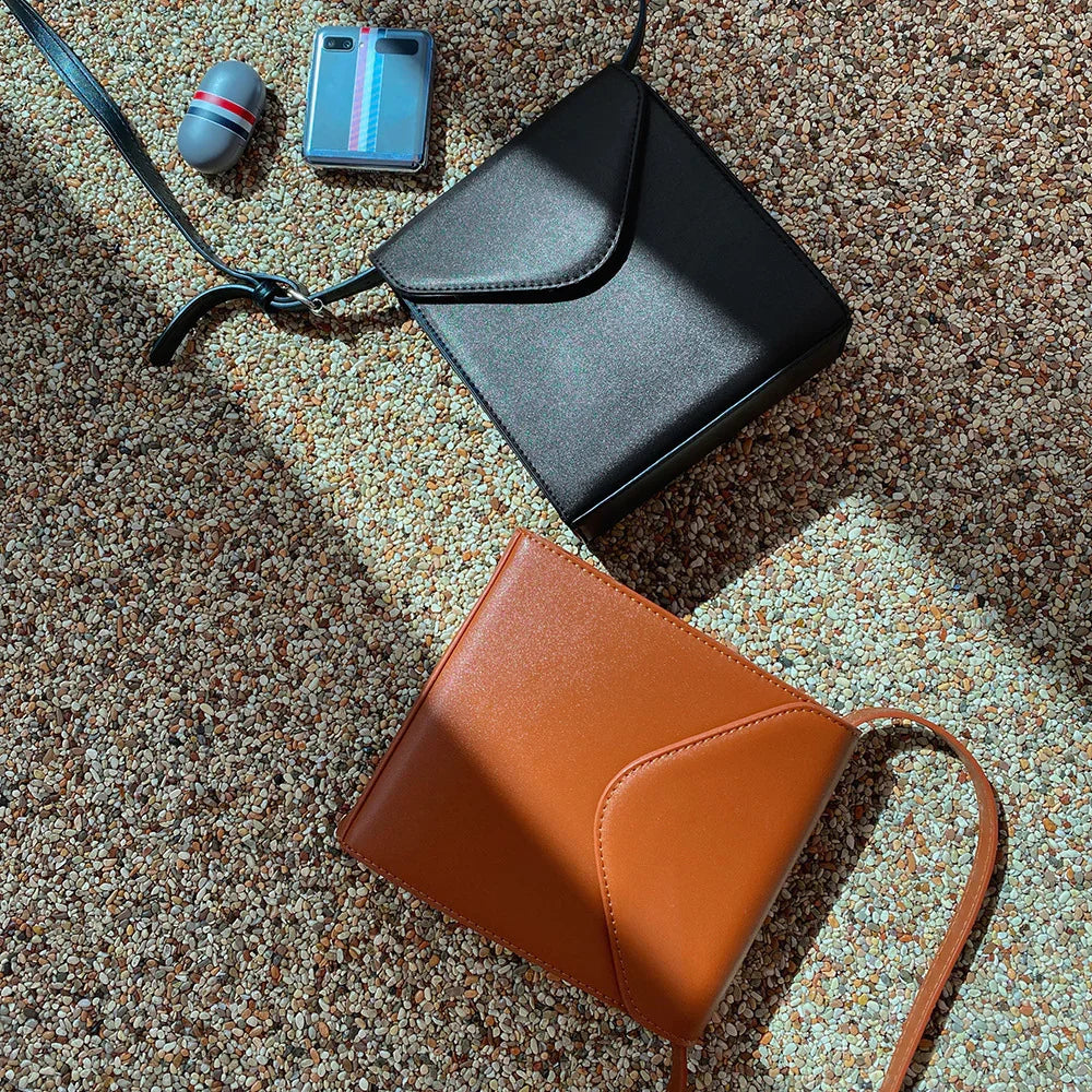 Two small leather crossbody bags, one black and one brown, on pebble-textured floor with sunlight