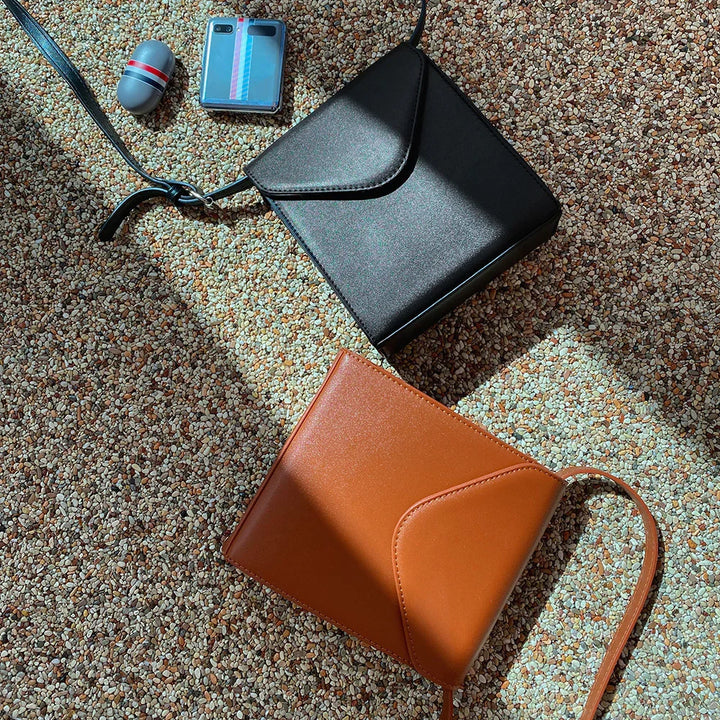 Two small leather crossbody bags, one black and one brown, on pebble-textured floor with sunlight