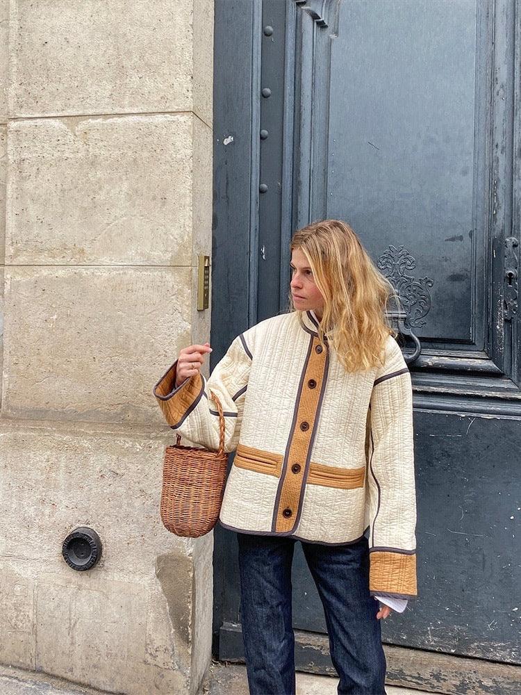 Woman wearing a cream and tan quilted jacket holding a wicker handbag, standing by a beige stone wall and dark gray door