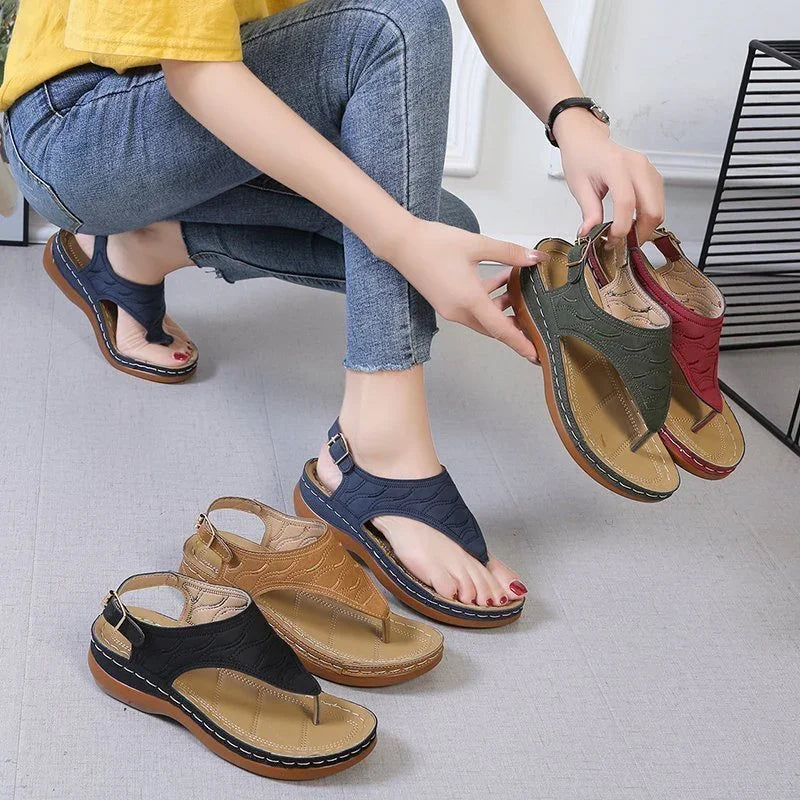 Woman wearing navy blue casual sandals, holding olive green and red sandals, with black and tan sandals on floor