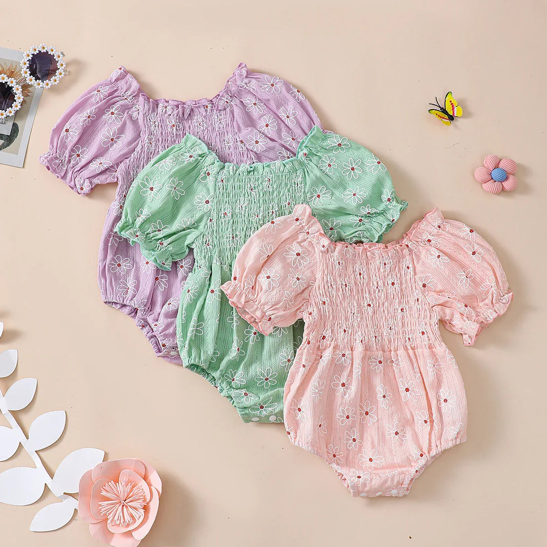 Three floral baby rompers in purple, green, and pink with puff sleeves and smocked bodices