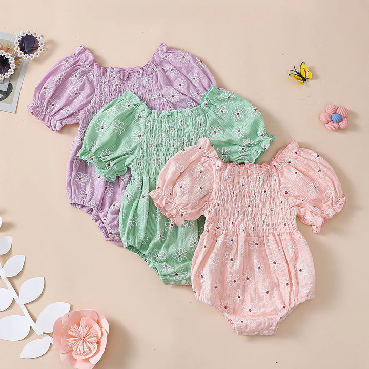 Three floral baby rompers in purple, green, and pink with puff sleeves and smocked bodices