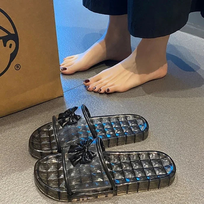 Black transparent slide sandals with bow detail beside bare feet on gray floor