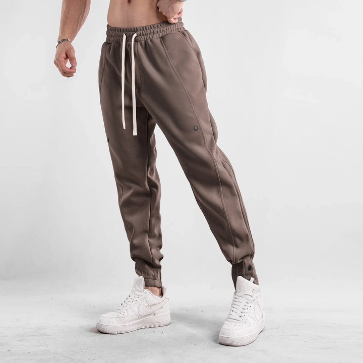 Man wearing brown jogger pants with white drawstrings and white designer sneakers on gray floor