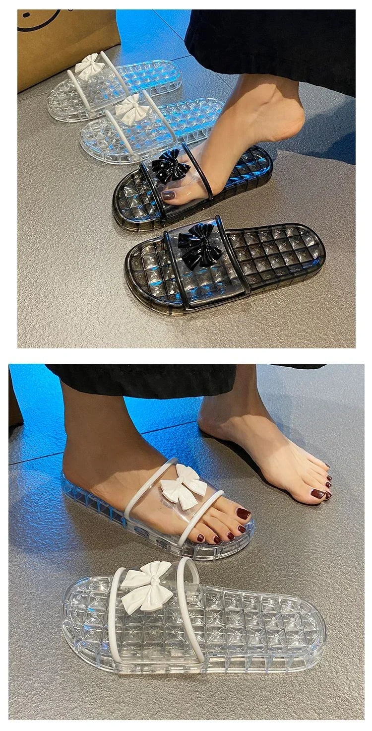 Clear plastic slide sandals with white and black bows, worn by feet with dark nail polish on a textured floor