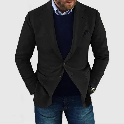 Man wearing black blazer over navy sweater and blue jeans on light gray background