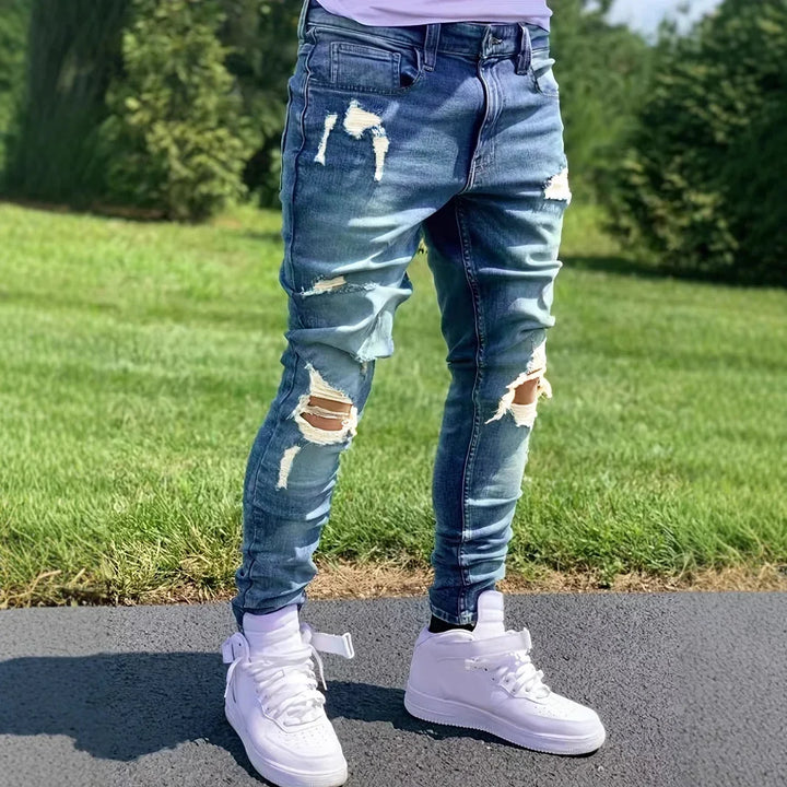 Men's ripped skinny blue jeans paired with white high-top sneakers on asphalt with green grass background
