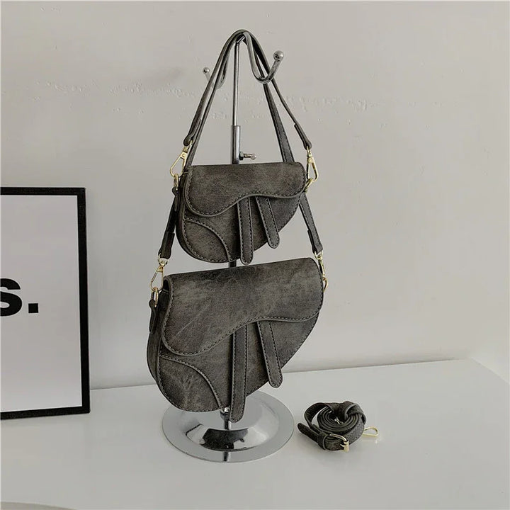 Two gray leather saddle bags with adjustable straps displayed on a metal stand on a white surface