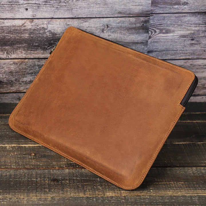 Brown leather laptop sleeve on rustic wooden surface with visible zipper and stitching