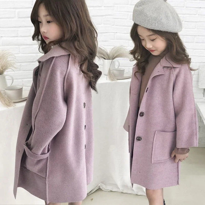 Two young girls modeling soft pink wool coats in a bright, modern room with white brick wall