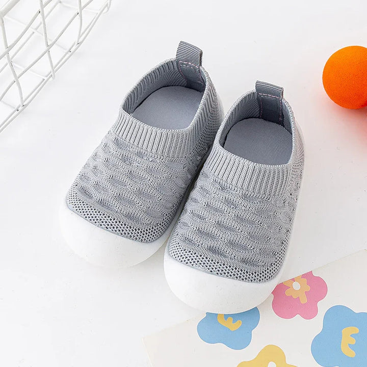 Pair of gray knitted baby shoes with white rubber soles on white surface with colorful floral paper and orange ball