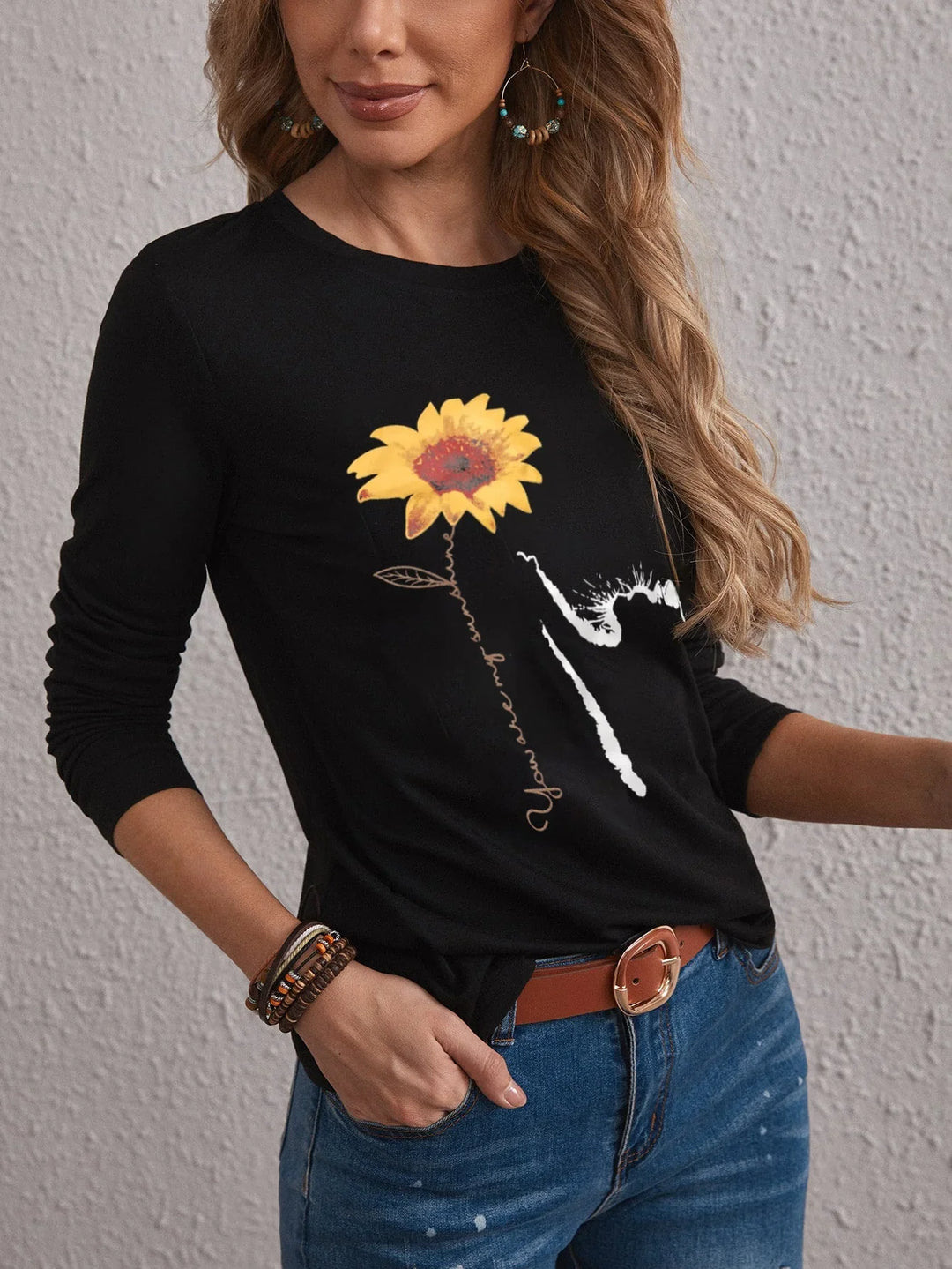Woman wearing black long sleeve top with yellow sunflower graphic and text, paired with blue jeans and accessories