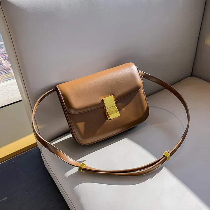 Sleek brown leather shoulder bag with gold clasp resting on light gray sofa under sunlight