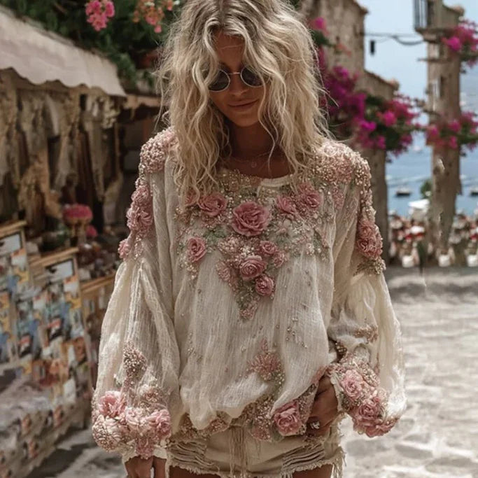 Blonde woman wearing embroidered floral blouse and sunglasses walking in sunlit street with flowers and sea view