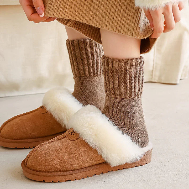 Person wearing brown shearling slippers with white fur lining and thick knitted socks indoors