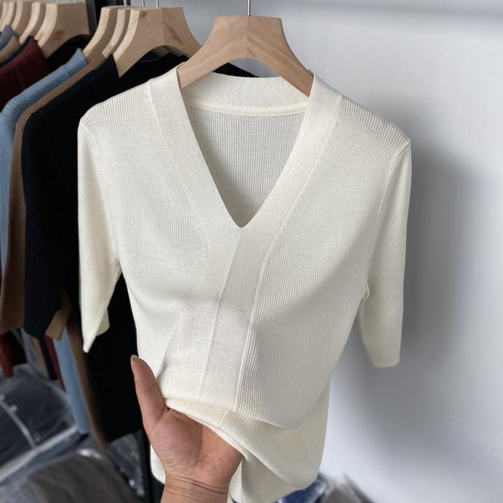 Hand holding a cream ribbed knit V-neck top with half sleeves on wooden hanger, clothing rack with assorted sweaters in background