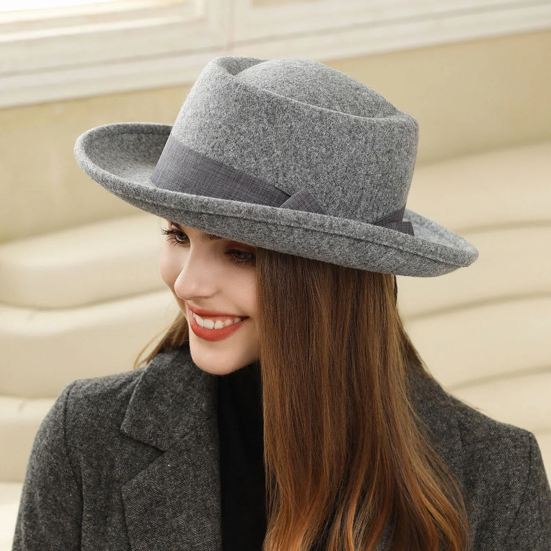 Smiling woman wearing a gray felt fedora hat and dark gray coat indoors