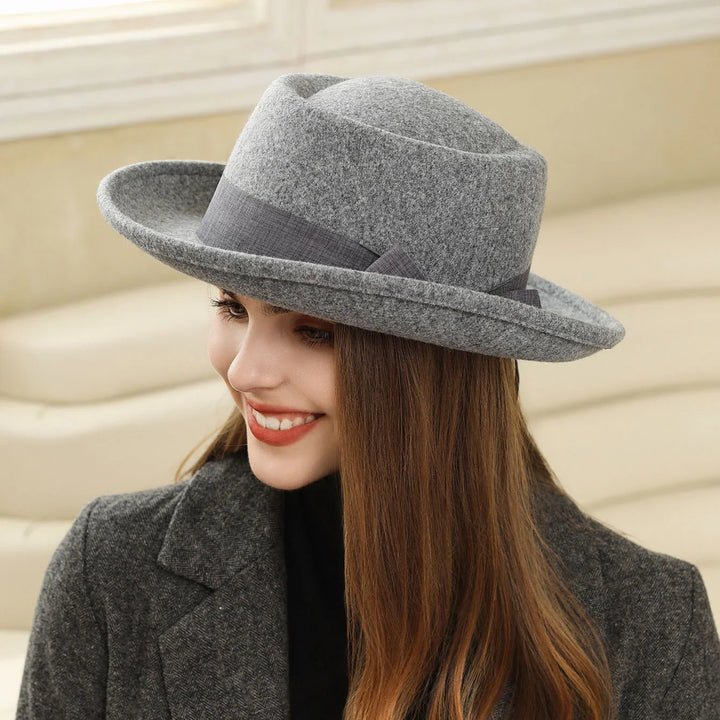 Smiling woman wearing a gray felt fedora hat and dark gray coat indoors