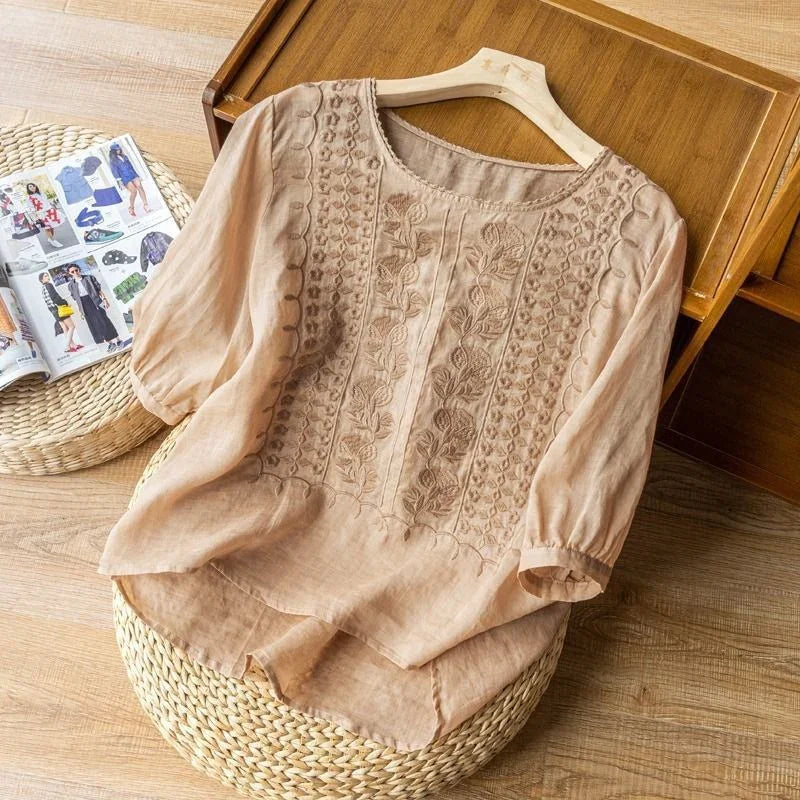 Beige linen blouse with floral embroidery on front, displayed on wooden hanger over wicker and wood background