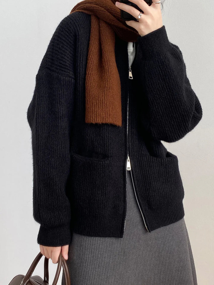 Person wearing black zip-up knit sweater, brown scarf, and gray ribbed skirt holding brown handbag