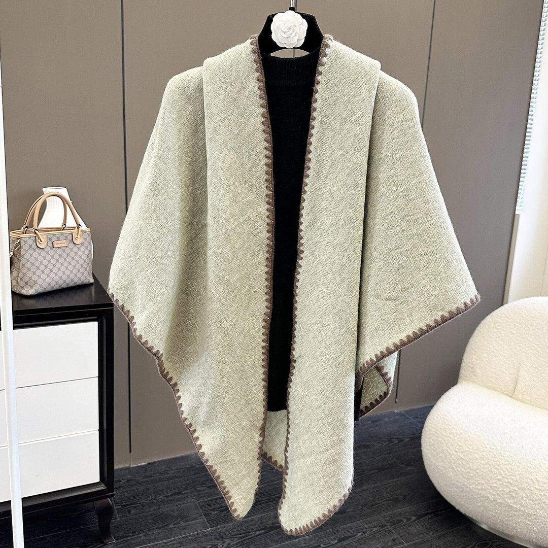 Beige wool poncho with brown trim displayed on black mannequin in modern room with handbag and white chair