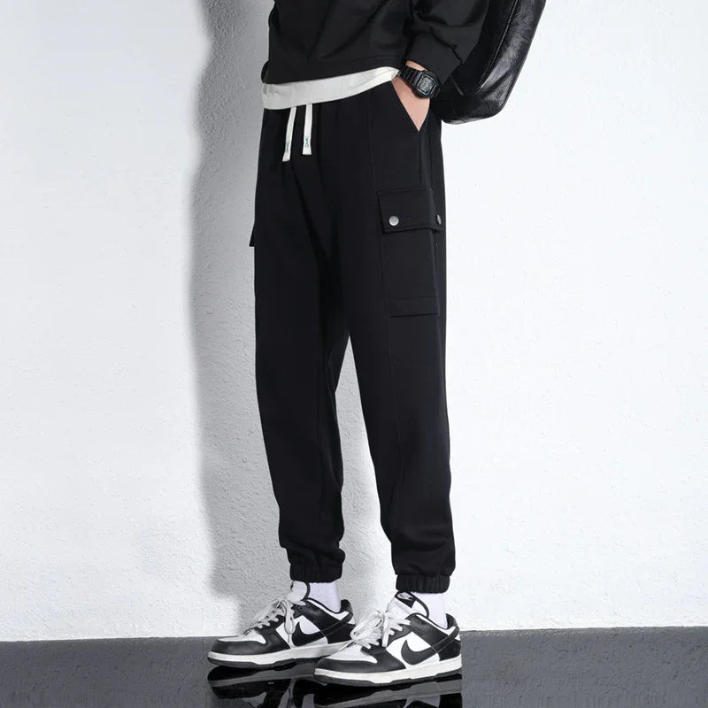 Man wearing black cargo jogger pants with elastic cuffs and black white sneakers against white wall