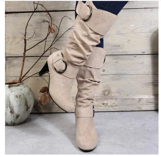 Beige slouchy knee-high boots with decorative buckle straps worn by person against wooden background