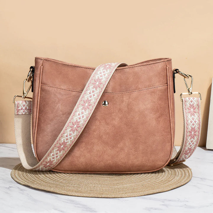 Brown faux leather crossbody bag with pink and white embroidered strap on marble surface