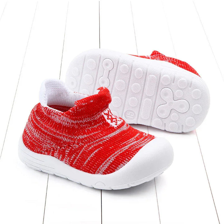 Red and white toddler knit slip-on shoes with flexible white rubber soles on white wooden floor