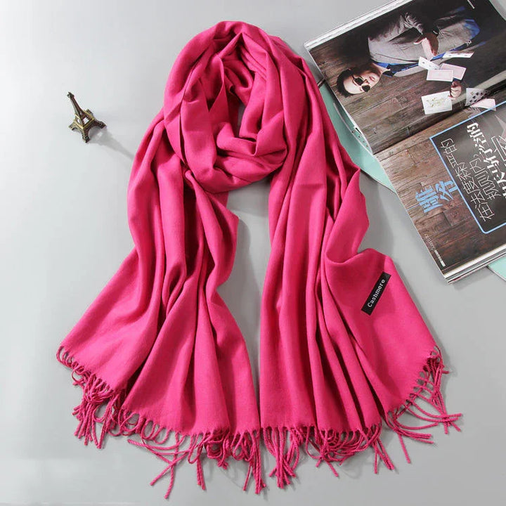 Bright pink cashmere scarf with fringed edges displayed on flat surface next to a miniature Eiffel Tower and an open magazine
