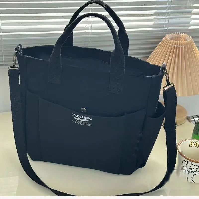 Black cloth bag with handles and adjustable shoulder strap on a white table near a lamp and cup