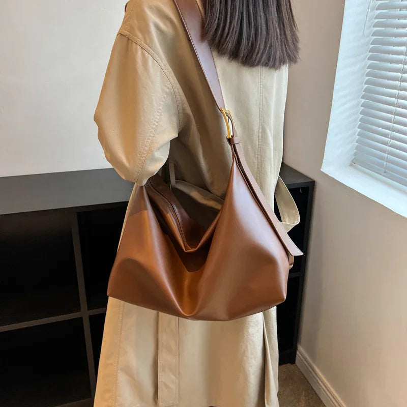 Brown leather shoulder bag worn by person in beige trench coat indoors near window blinds