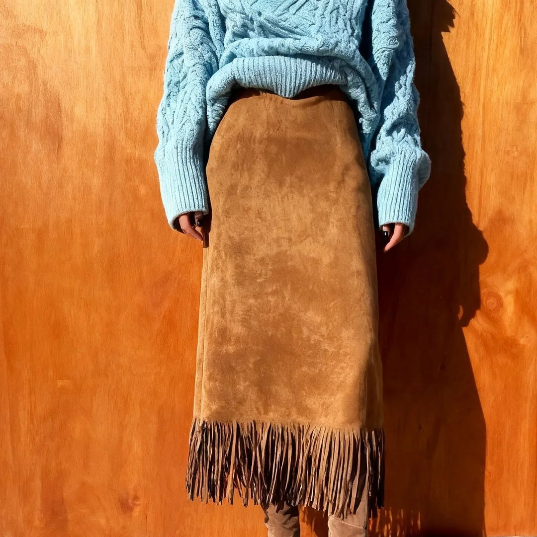 Woman wearing a light blue cable knit sweater and a brown suede fringe skirt against a wooden background