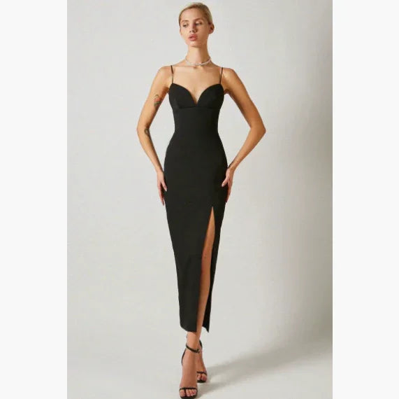 Woman in elegant black spaghetti strap dress with high thigh slit posing against plain backdrop