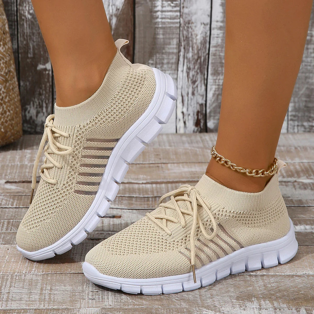 Beige knitted sock-style sneakers with white flexible soles worn with gold anklet on wooden floor