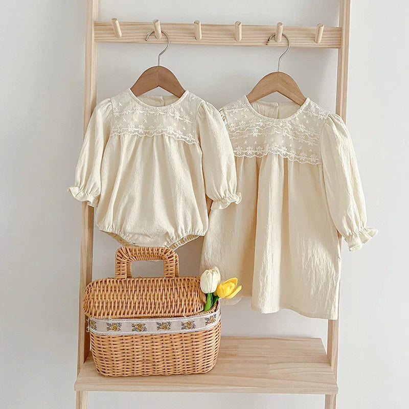 Beige baby romper and toddler dress with lace yoke on wooden hangers above wicker basket with tulips