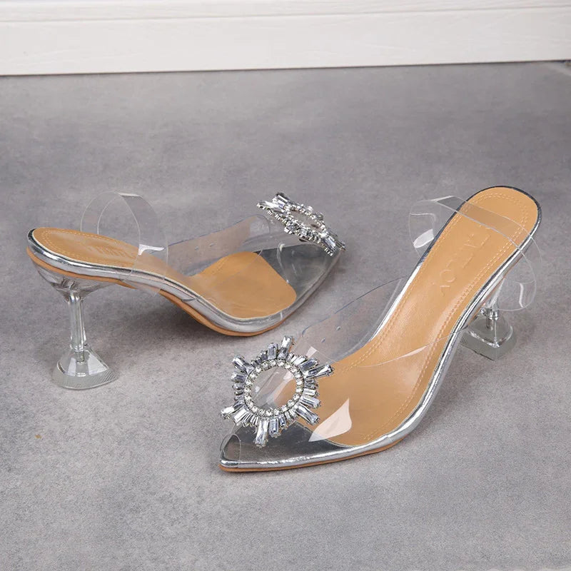 Transparent high-heeled mule shoes with jeweled circular embellishment and clear straps on gray surface
