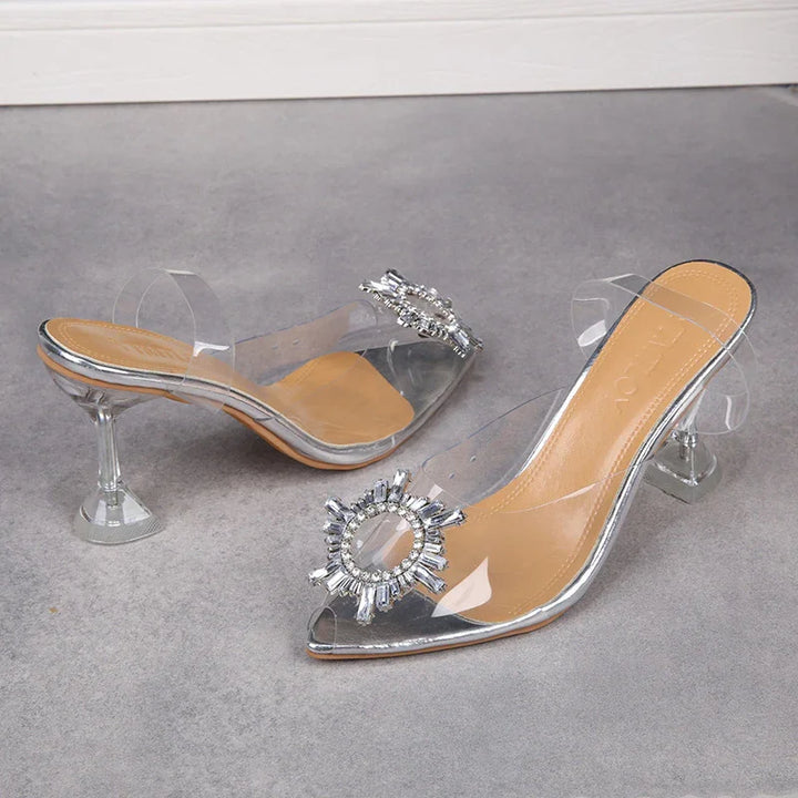 Transparent high-heeled mule shoes with jeweled circular embellishment and clear straps on gray surface