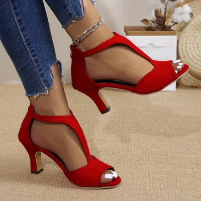 Close-up of feet wearing red high-heeled open-toe shoes with ankle strap and chain anklet, paired with frayed hem jeans on beige carpet