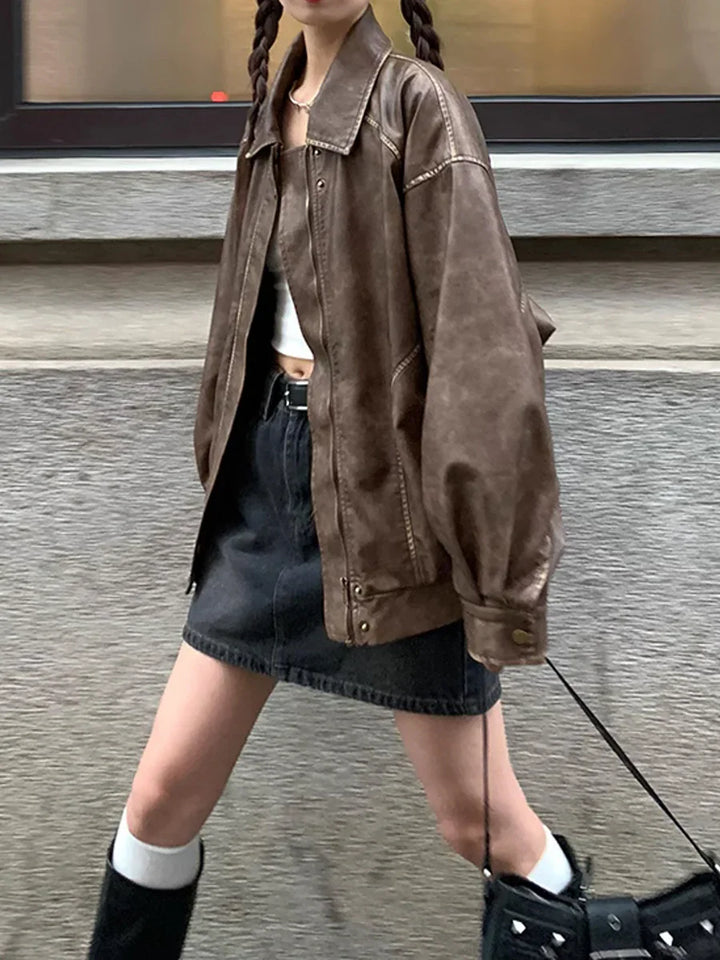 Young woman wearing oversized brown leather jacket, black denim skirt, white crop top, and black boots walking outdoors
