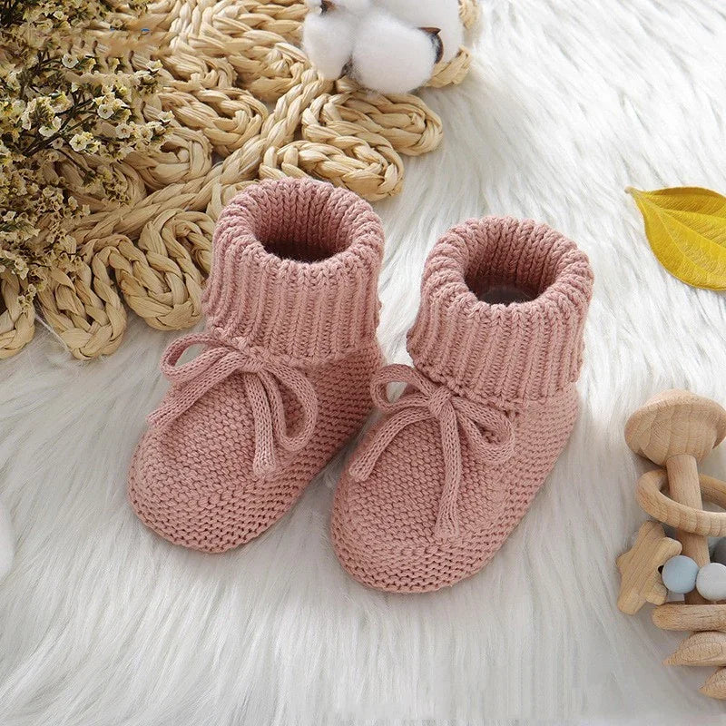 Pair of pink knitted baby booties with ties on white fluffy surface, surrounded by dried flowers and wooden toys