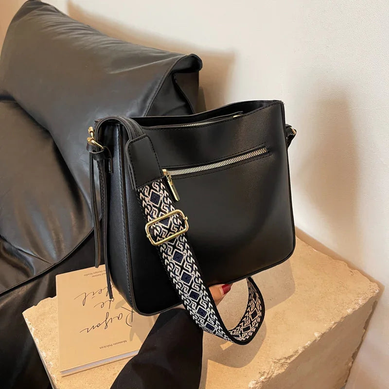 Black leather shoulder bag with patterned adjustable strap and front zipper pocket on stone surface