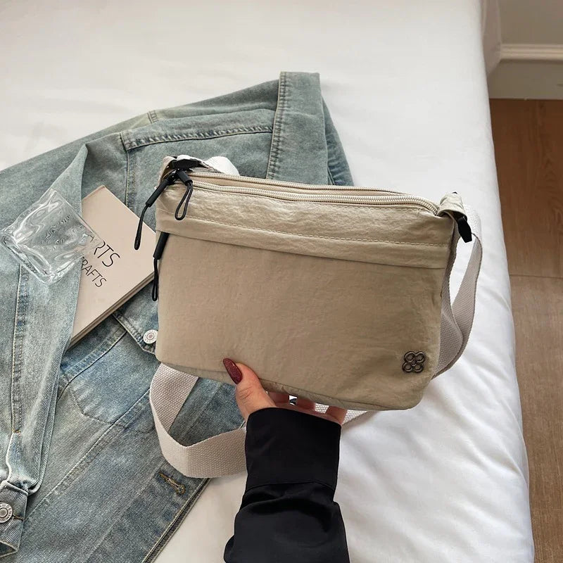Beige crossbody bag held over denim jacket and book on white bed with wooden floor