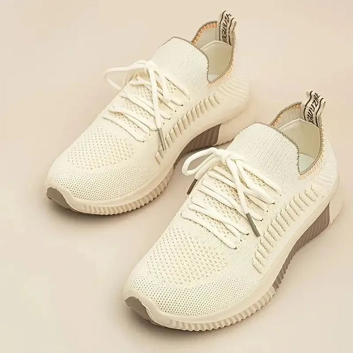 Pair of white knit athletic sneakers with textured soles and pull tabs on a beige background