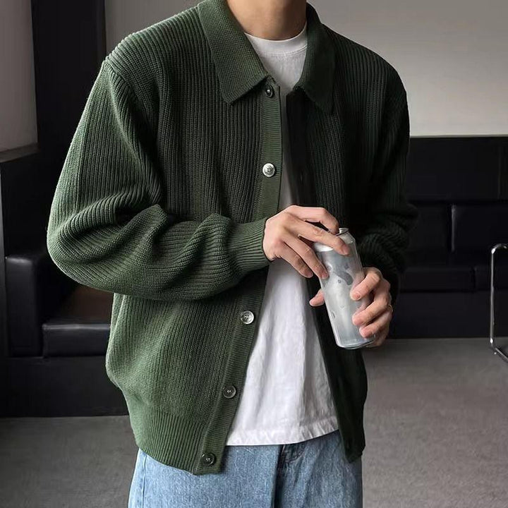 Man wearing dark green ribbed cardigan over white t-shirt and light blue jeans holding a silver can indoors